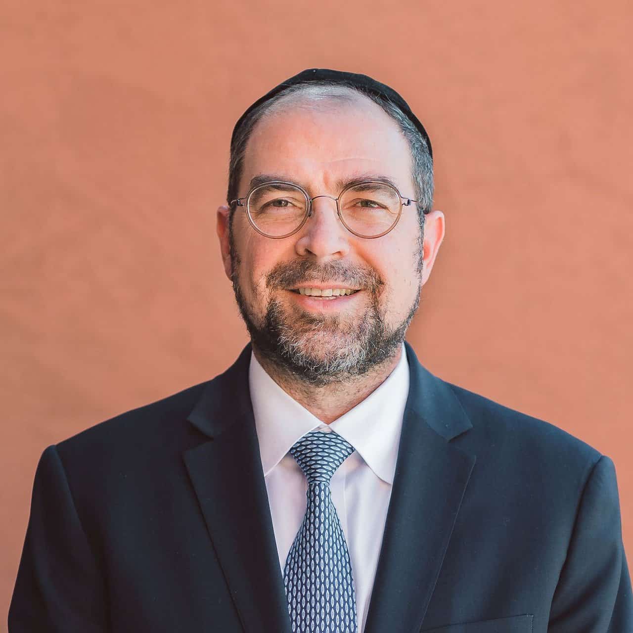 Professional man in business suit with glasses smiling in front of a warm background.