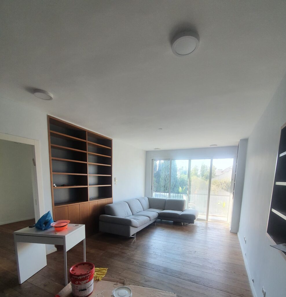 Bright living room with large windows, modern gray sectional sofa, and wooden floor. Empty bookshelf and sliding glass door leading to balcony.