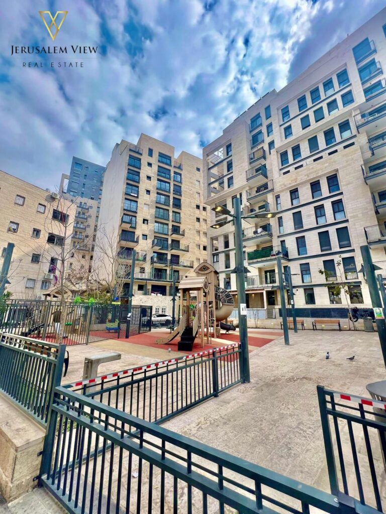 Modern apartment complex with a children’s playground and open courtyard, under a cloudy sky in Jerusalem.