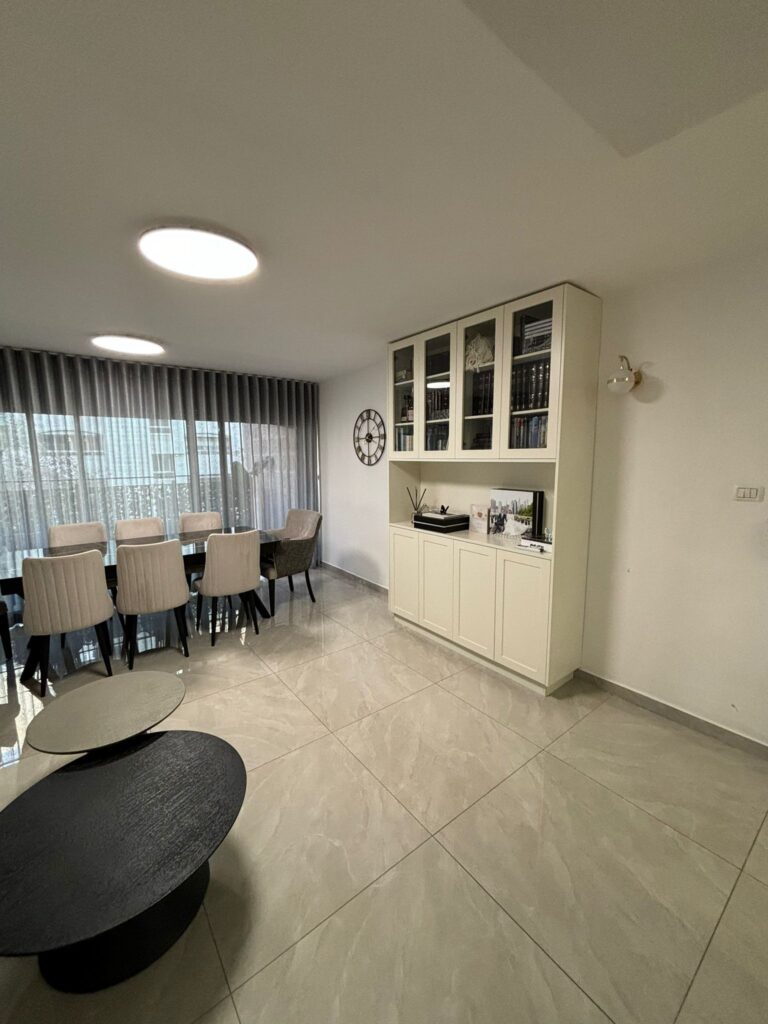 Modern dining room with large window and elegant furniture, featuring a sleek black coffee table and white cabinetry.