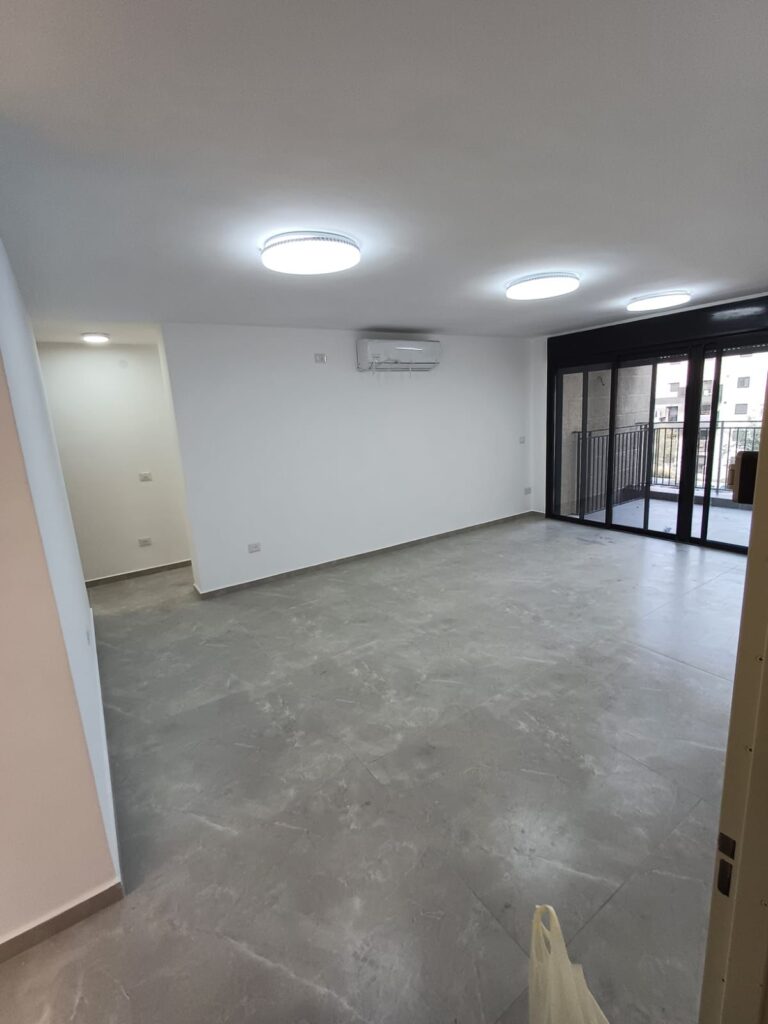 Modern living room with large glass sliding doors and balcony access, bright LED ceiling lights, white walls.