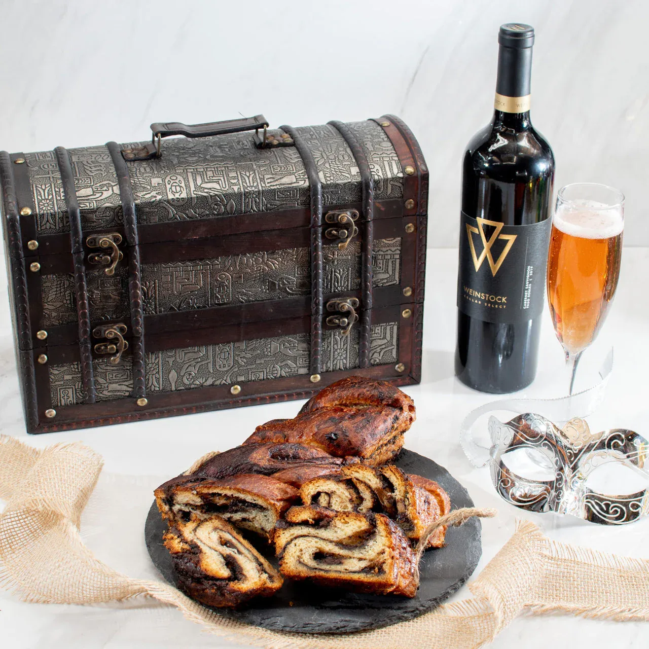 Vintage wine and cake set with masquerade mask, on a white table with a decorative chest.