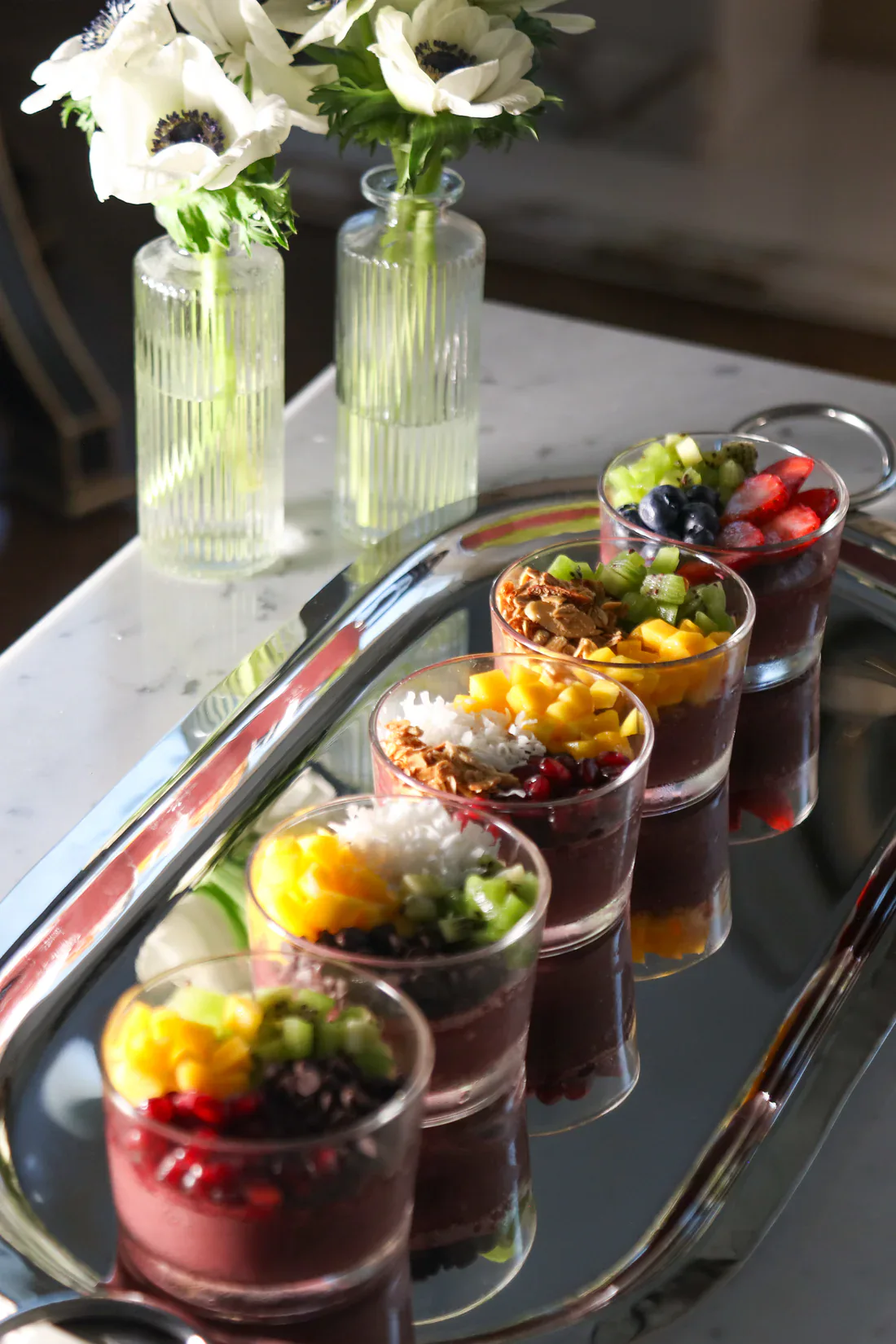 Vibrant fruit parfaits in small glasses arranged on a tray with fresh white flowers in the backgroun.