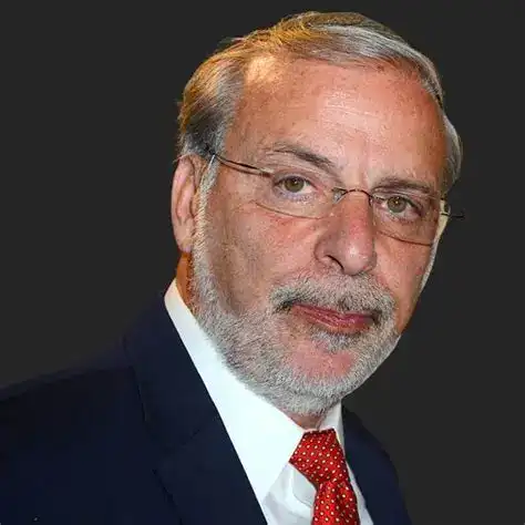 Elderly man with gray hair, beard, glasses, dressed in a formal suit and red tie.