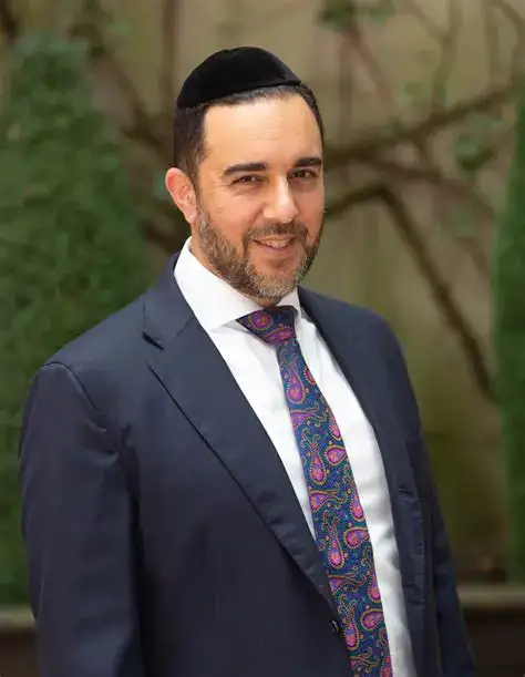 Businessman in formal suit smiling outdoors with greenery background.