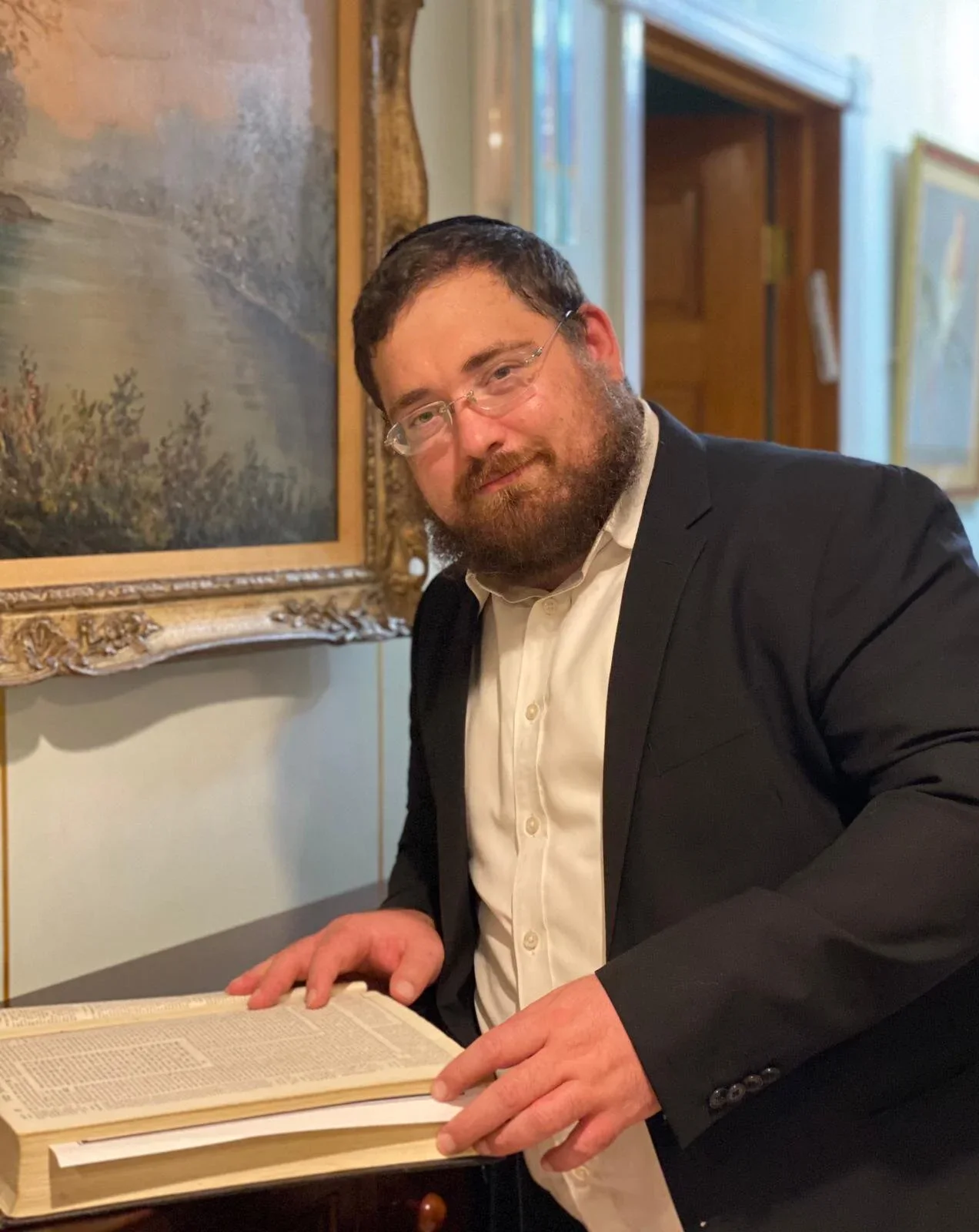 Man in formal attire reading a large book in an art gallery or museum setting.